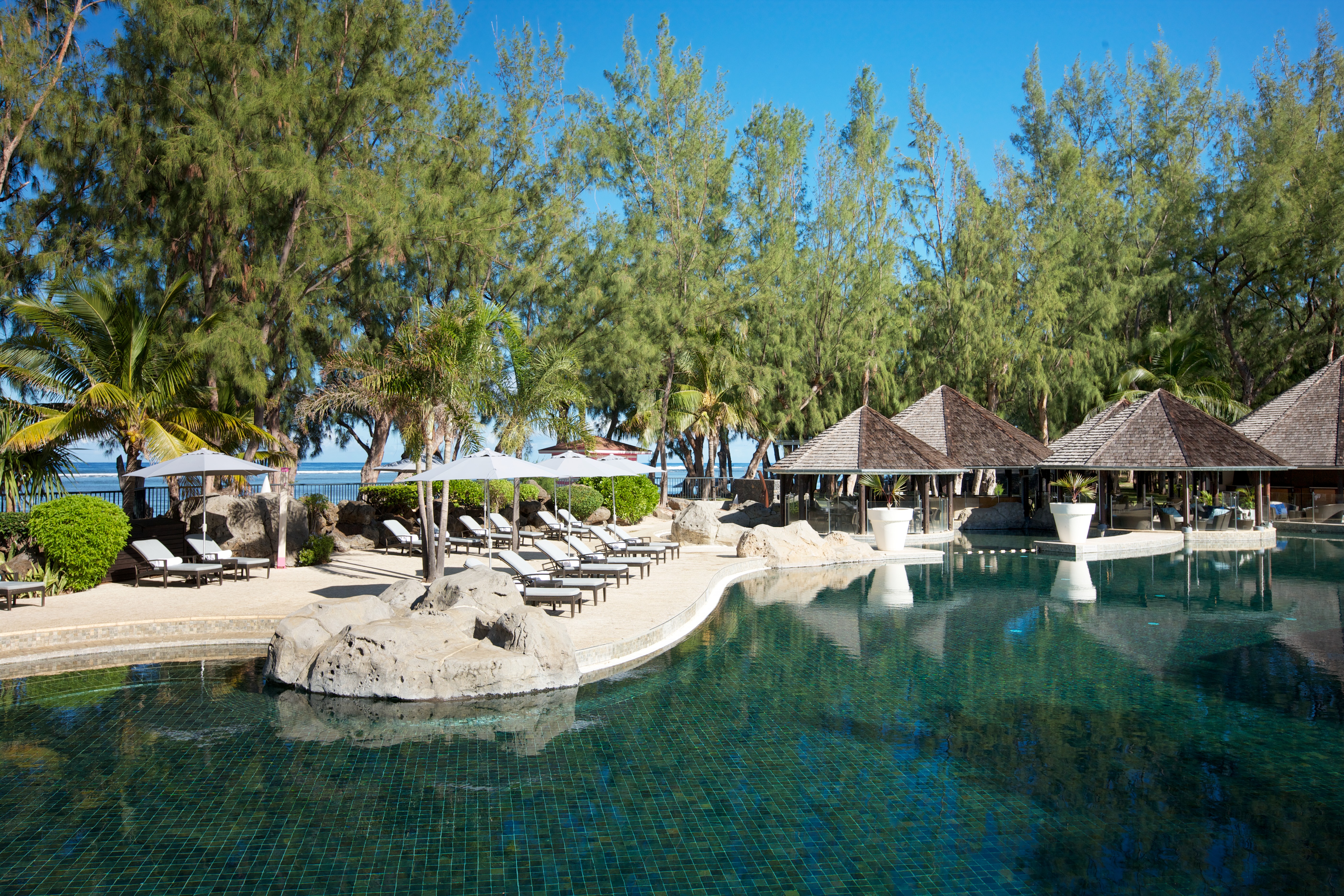 The alluring blue waters of the pool lie calm beneath trees and loungers offering shade at LUX* Saint Gilles Reunion